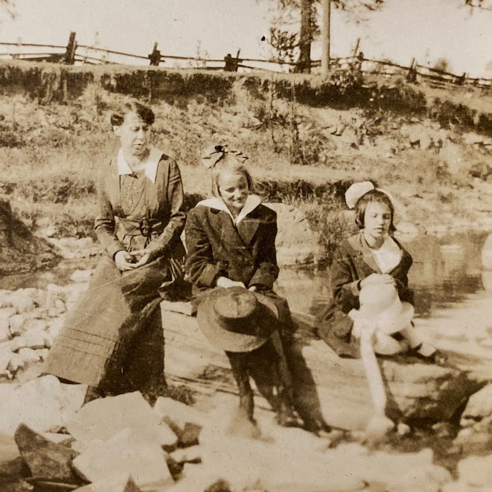 Two young girls and their aunt sit on rocks in Mimico Creek, about 1919