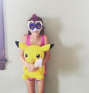 Little girl in pink shorts, tank top, and Mardi Gras-style mask, holding a huge yellow Pikachu